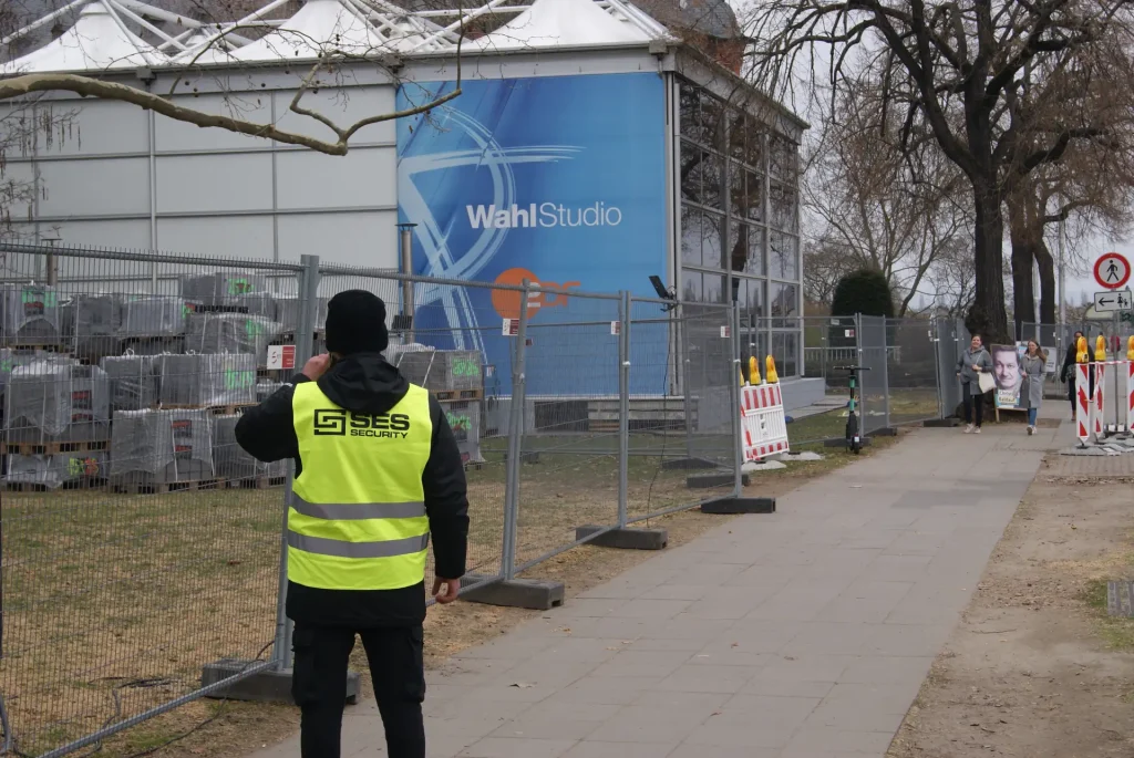EIn Mann mit schwarzer Mütze und schwarzer Kleidung und einer gelben Warnweste mit dem Schriftzug SES Group bewacht gerade ein Areal für Sicherheitsdienst Neustadt